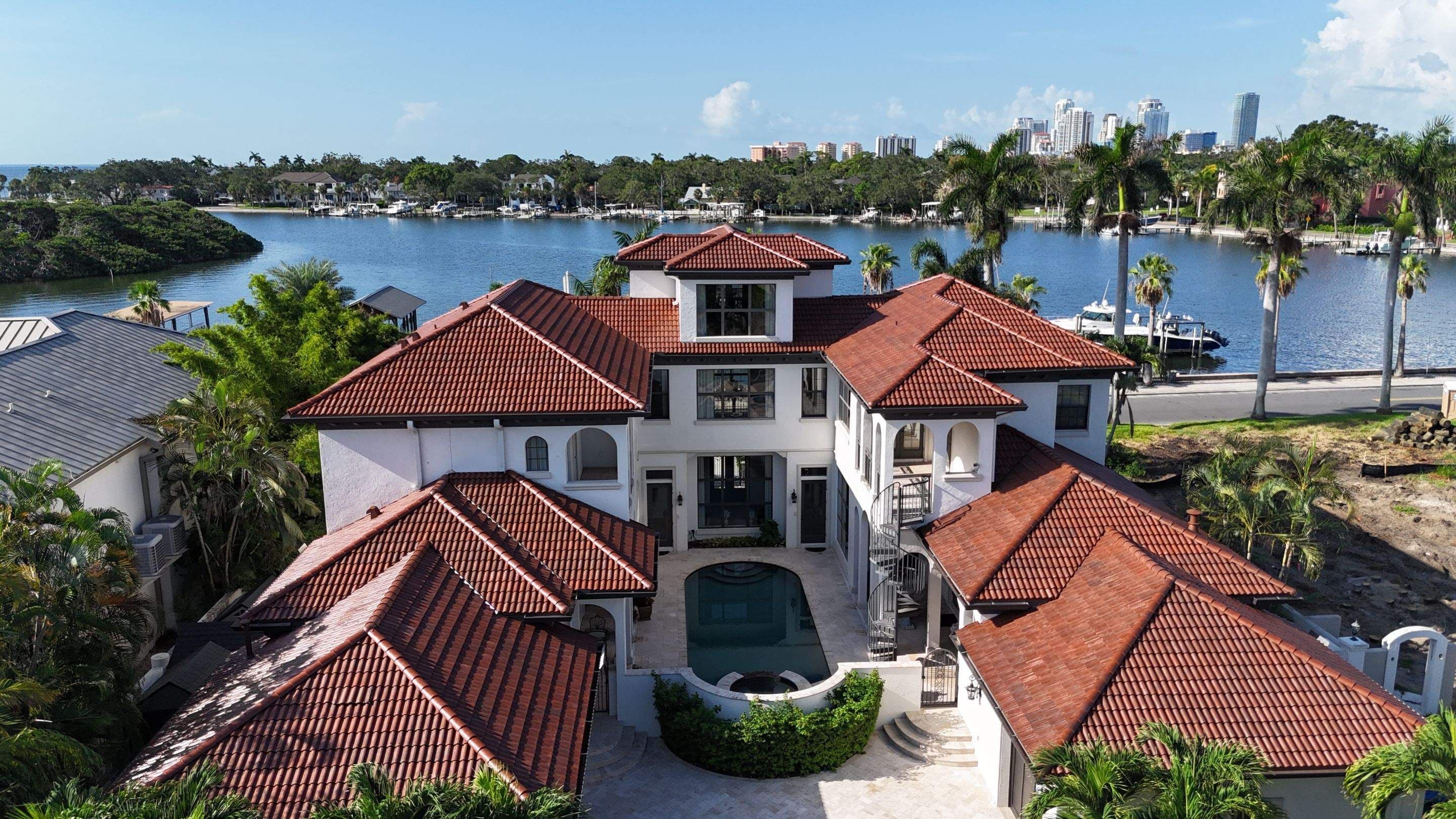 Red Spanish barrel tile roofs on a white waterfront home with a courtyard pool, the St. Petersburg skyline across the water, seen from a drone